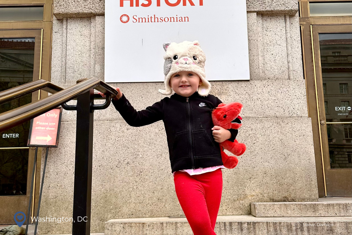 Skip Generation Travel Benefits (2) child in front on smithsonian museum wearing a snow cap and carrying a red stuffed dinosaur